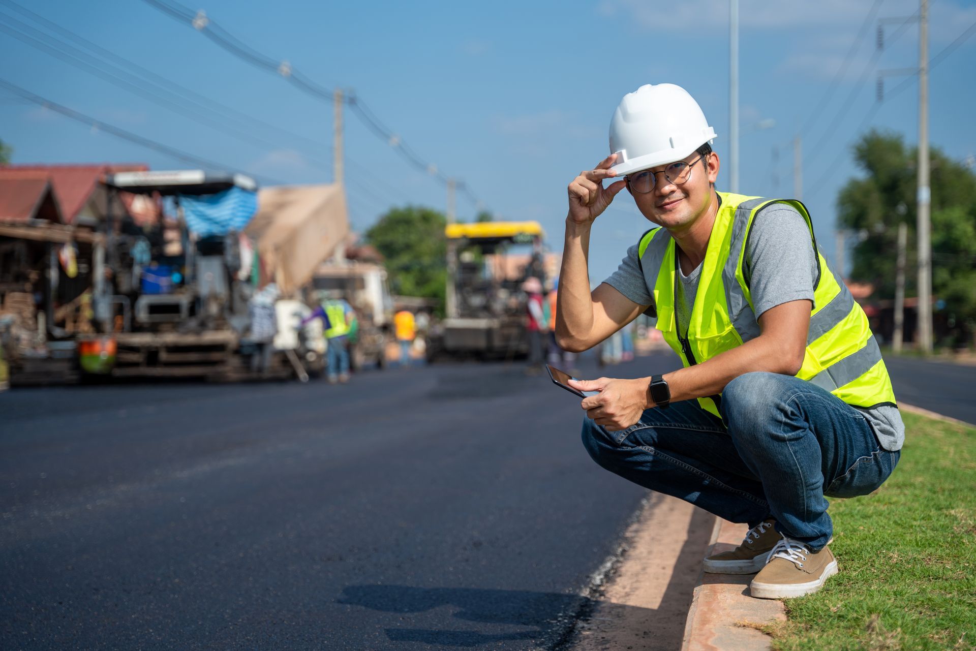 Construction worker kneels at roadside, wearing hardhat and safety vest, with paving equipment in the background. Construction worker kneels at roadside, wearing hardhat and safety vest, with paving equipment in the background.