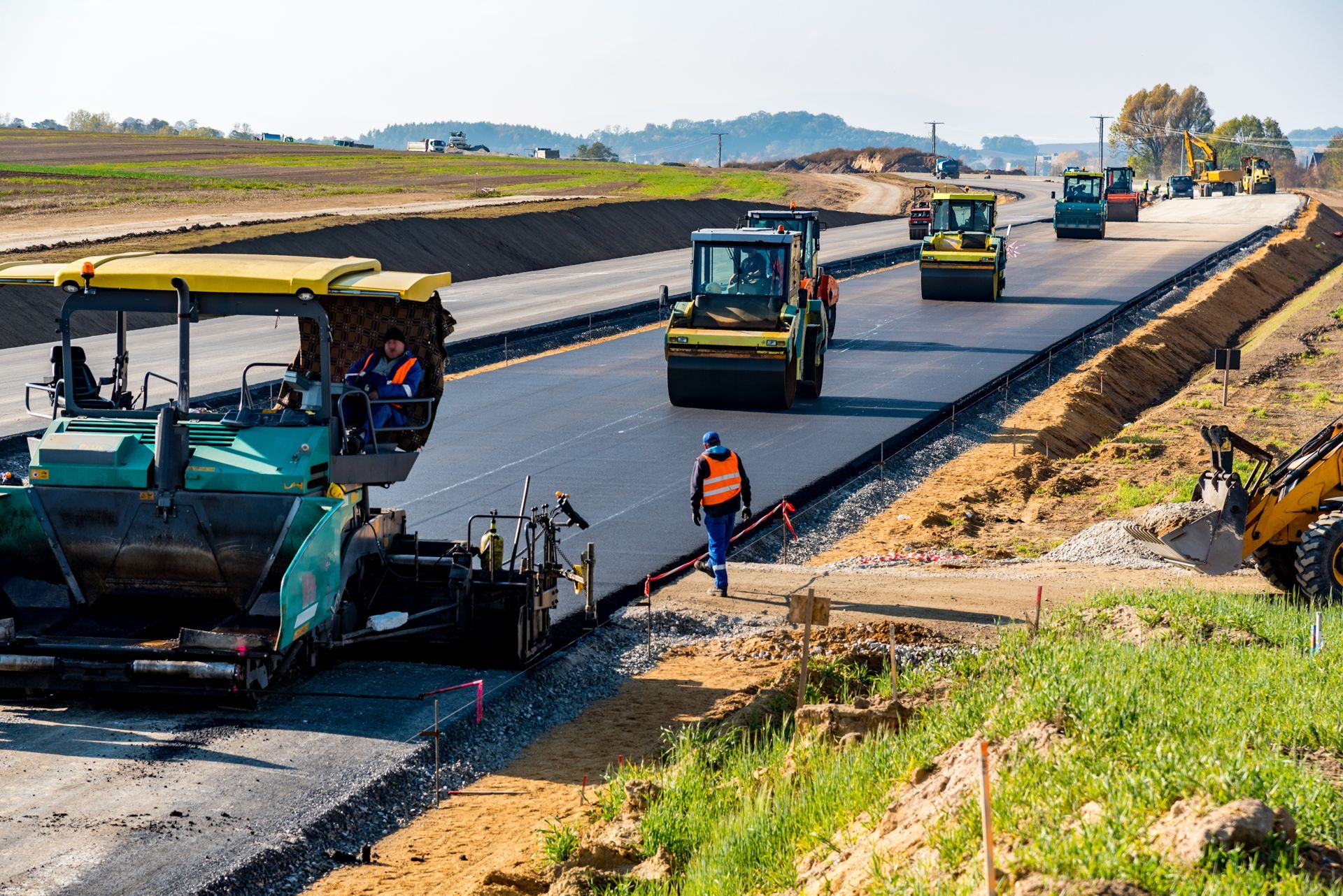 Road construction: Asphalt paver and rollers on a newly paved road, workers in orange vests, sunny day. Road construction: Asphalt paver and rollers on a newly paved road, workers in orange vests, sunny day.
