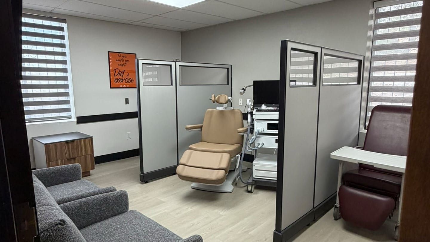 Small medical exam room with a tan exam chair, TMS therapy device, privacy partitions, and gray seating.