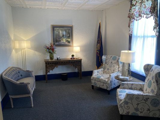 Waiting room with blue floral chairs, a sofa, and a table with a painting and flowers. A flag stands in the corner.