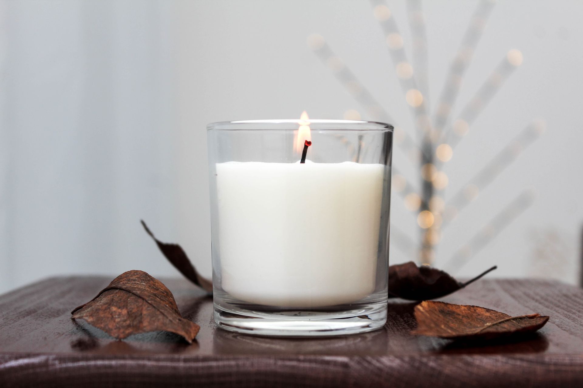 Lit white candle in a clear glass on a wooden surface, with dried leaves and string lights in the background.