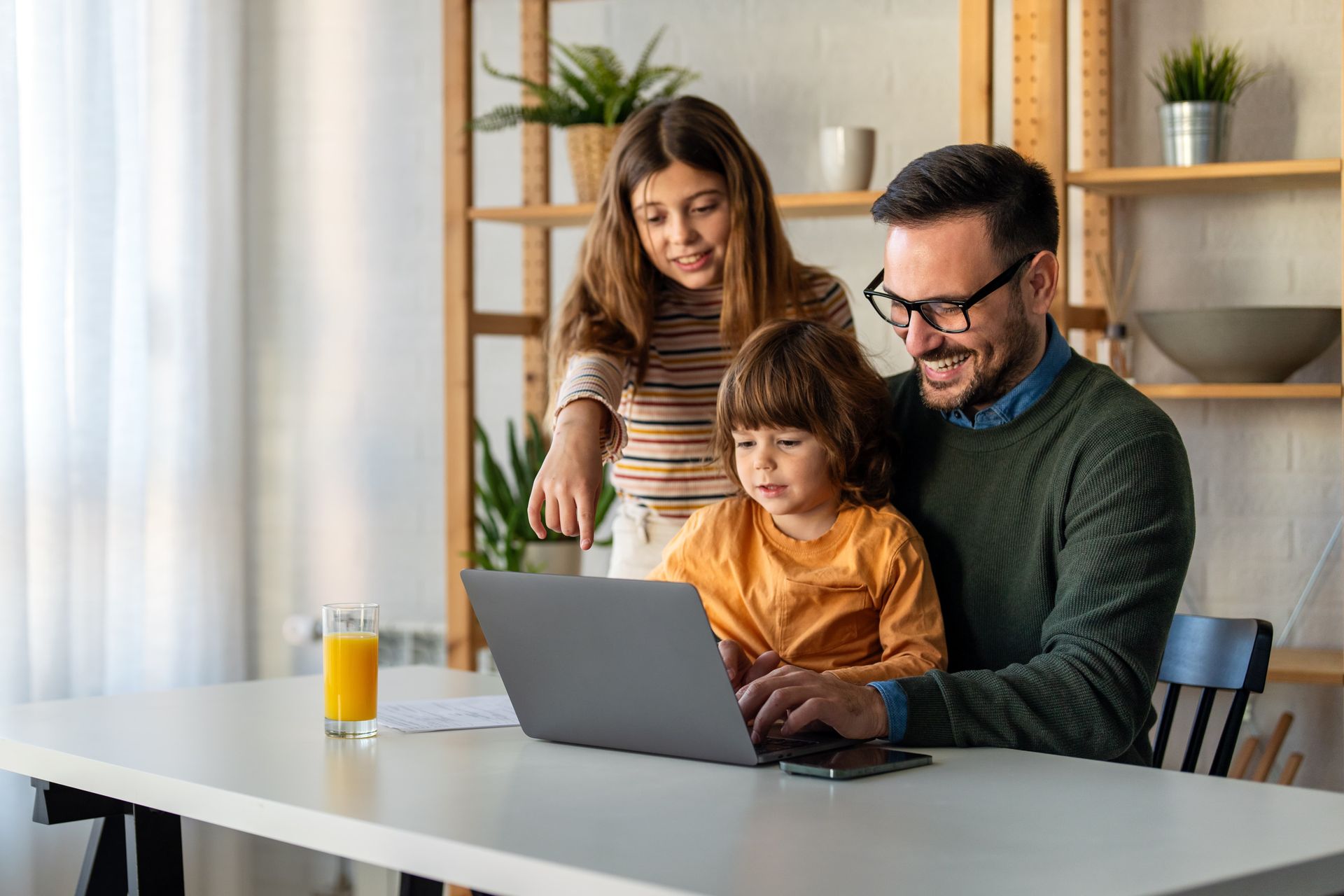 Father and two children smiling at laptop. Woman points at screen. Juice glass on table.
