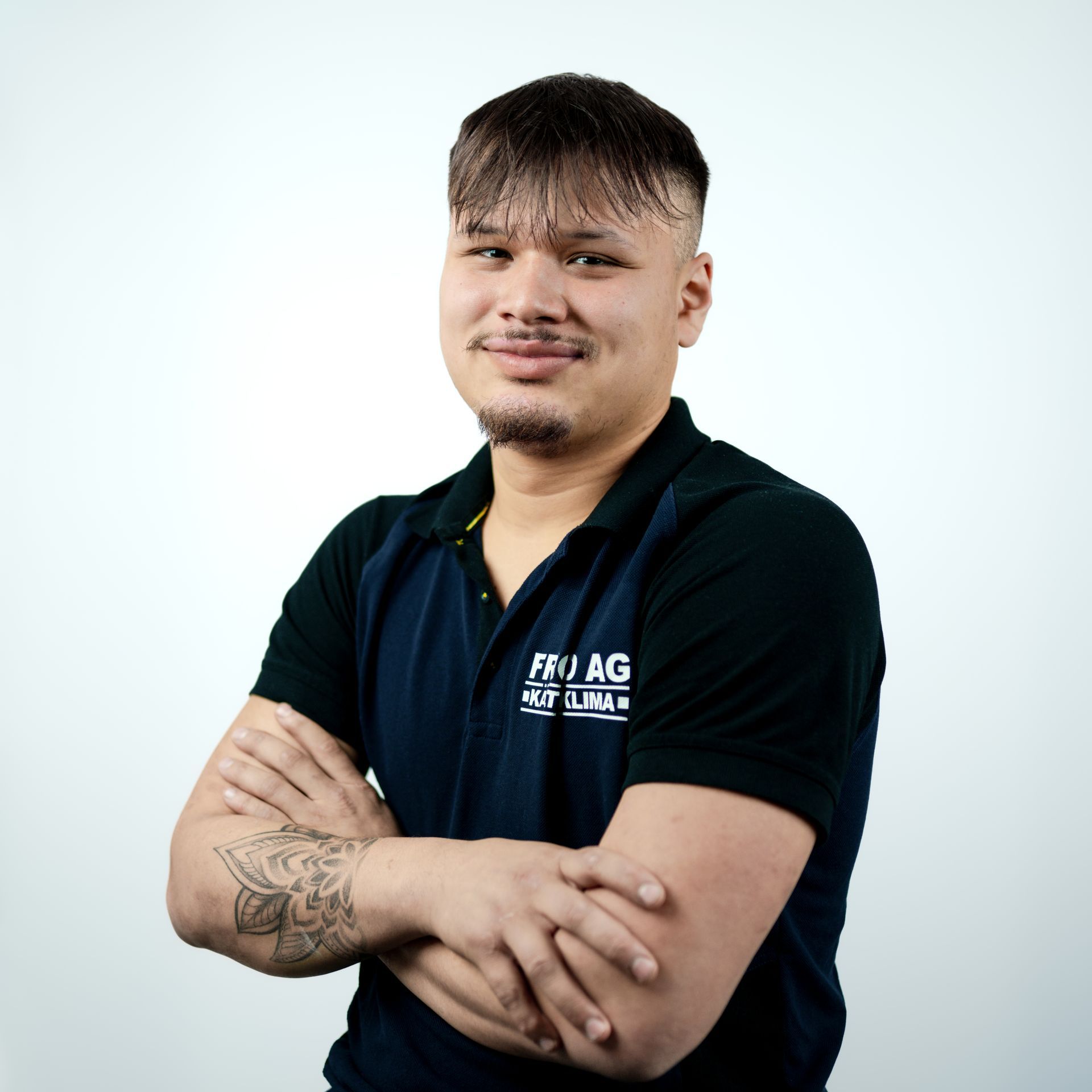 Mann mit verschränkten Armen, der ein dunkelblaues Poloshirt trägt, lächelt vor weißem Hintergrund.