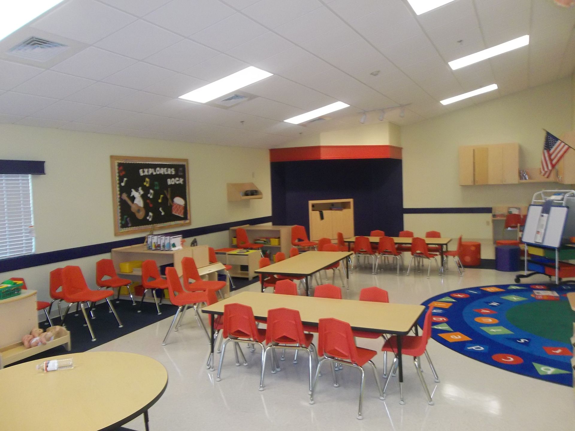 Classroom with red chairs and tables; colorful rug; walls are beige, red, and blue.