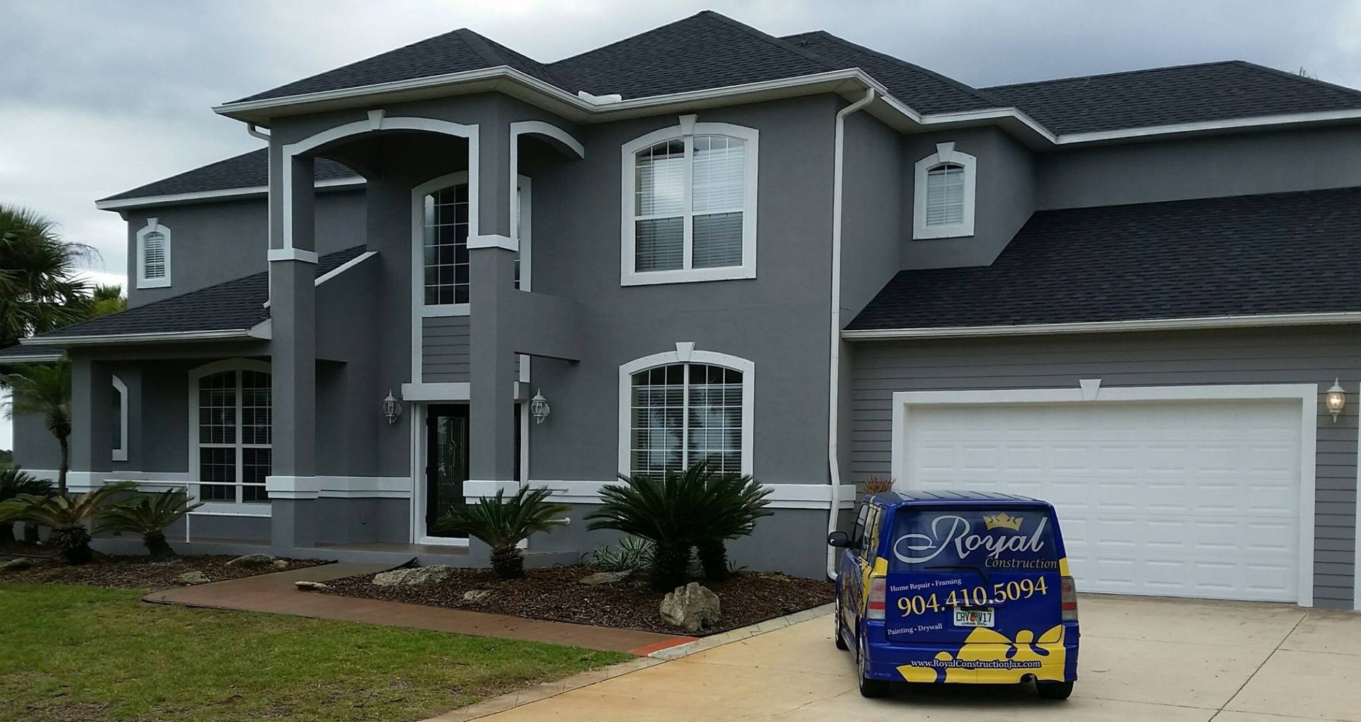 Gray stucco house with white trim, black roof. A blue company van is parked in front of the garage.