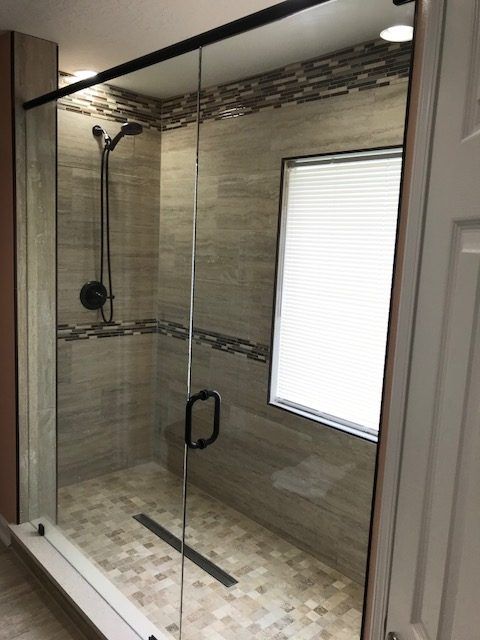 Shower stall with glass doors, black hardware, beige tile, small window with blinds.