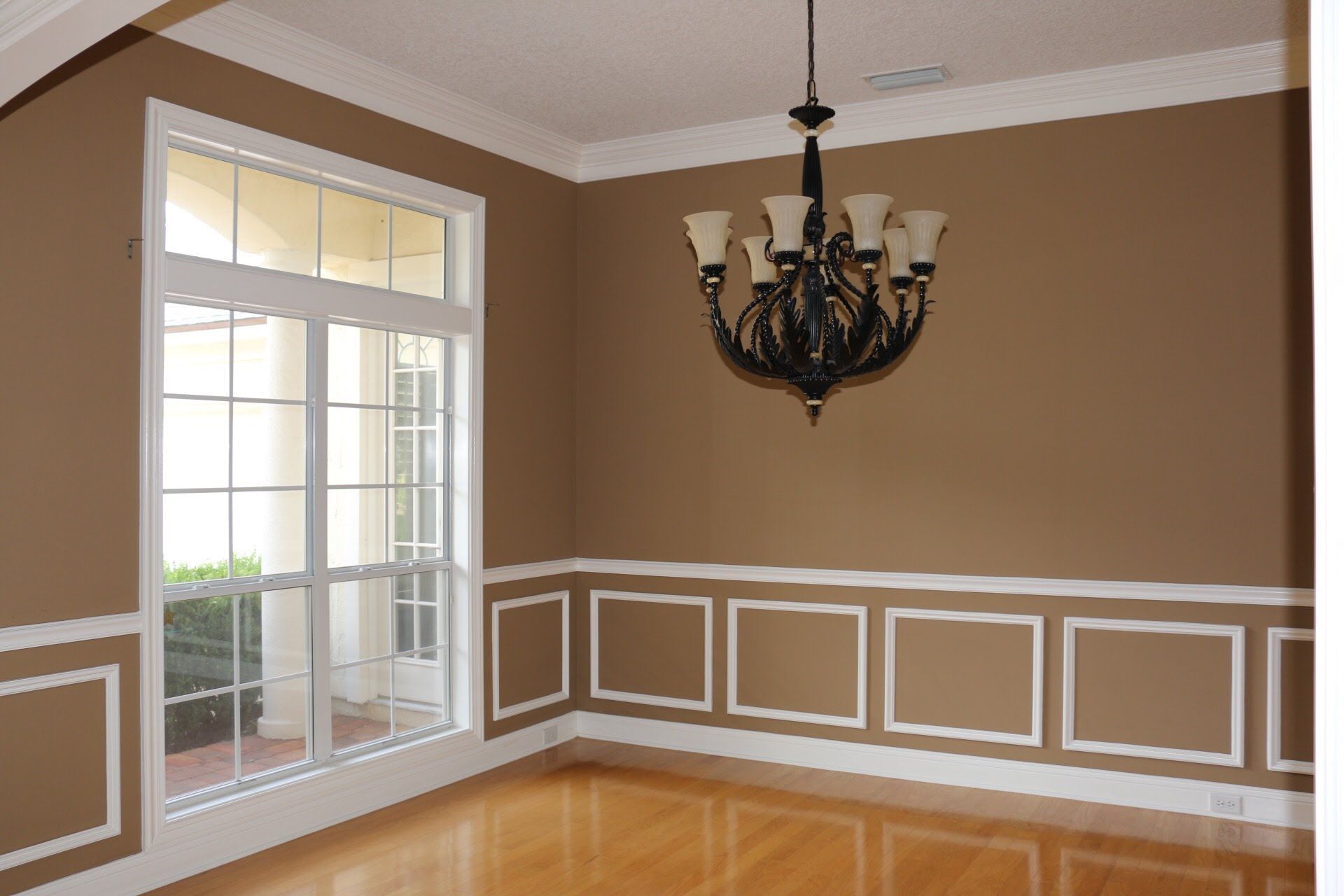 Empty dining room with brown walls, white trim, large window, chandelier, and hardwood floor.