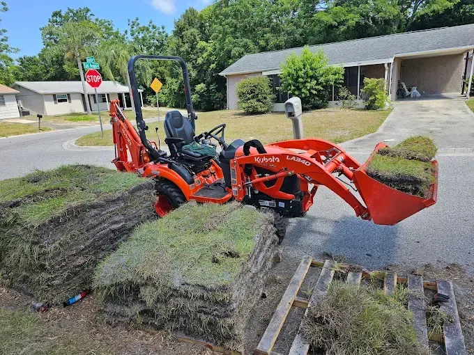 Orange tractor lifting sod by a suburban curb with two workers in a residential neighborhood