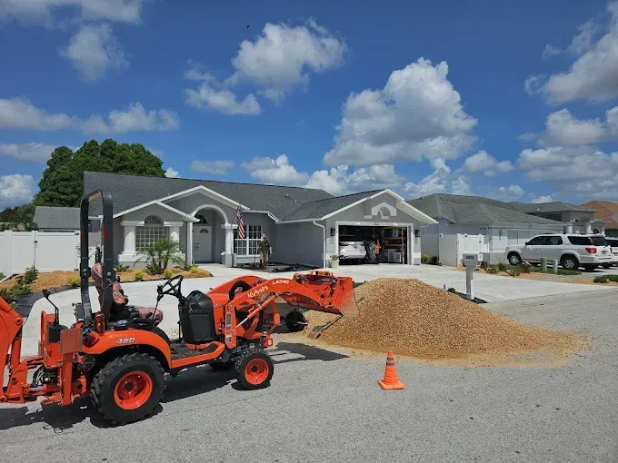 Orange tractor in front of a house, with gravel pile and orange cone on a driveway under a blue sky