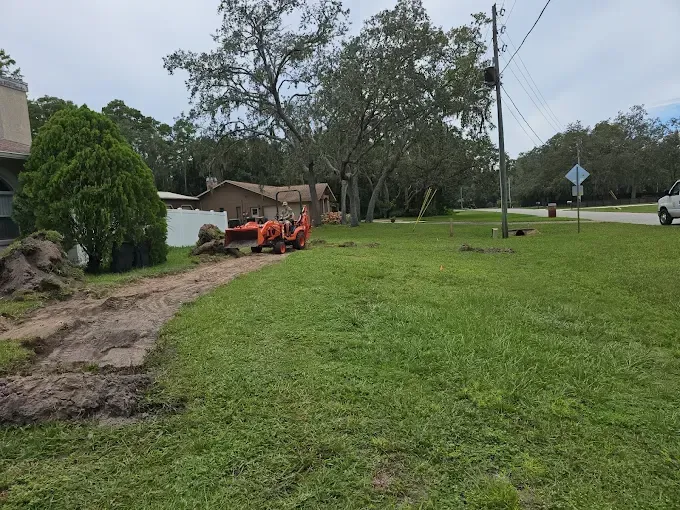 Orange backhoe digging along a grassy roadside near houses and trees