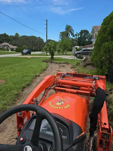 View from orange tractor driving along a grassy roadside near houses and trees