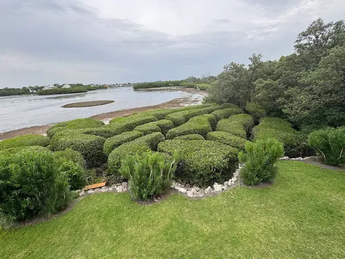 Manicured green hedges beside a grassy waterfront with trees and calm water under a cloudy sky