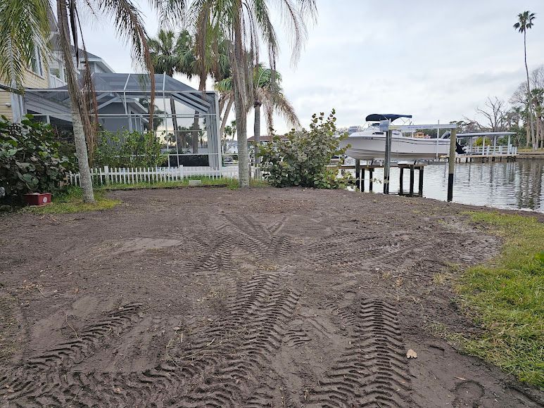 Sandy waterfront lot with palm trees, a small dock, and a building across the water