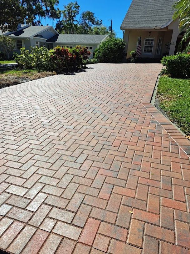Brick driveway leading to a house and garage, bordered by shrubs and grass under a sunny blue sky