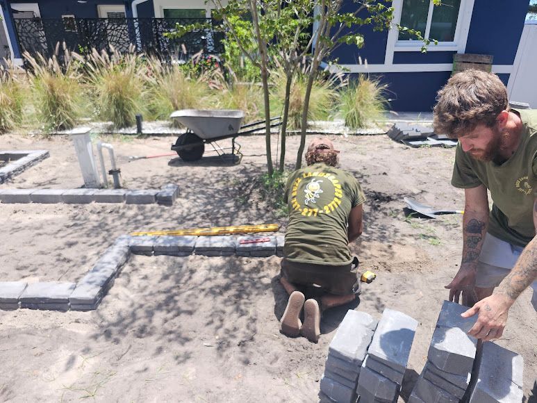Workers kneeling in a landscaped area, arranging paving stones near a building with a wheelbarrow nearby.
