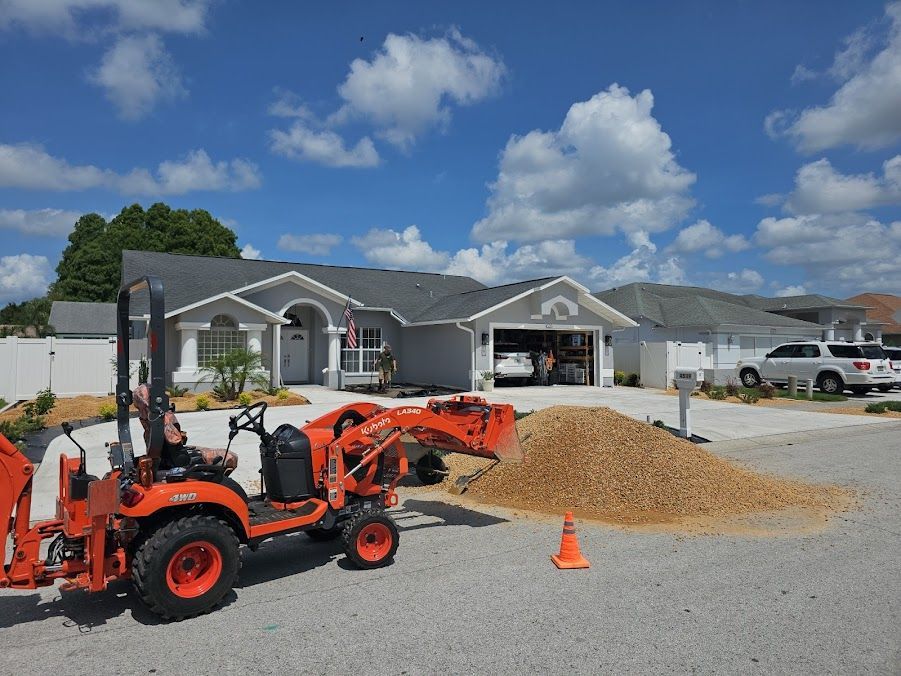 Orange tractor loader by a gravel pile outside a gray house on a sunny day