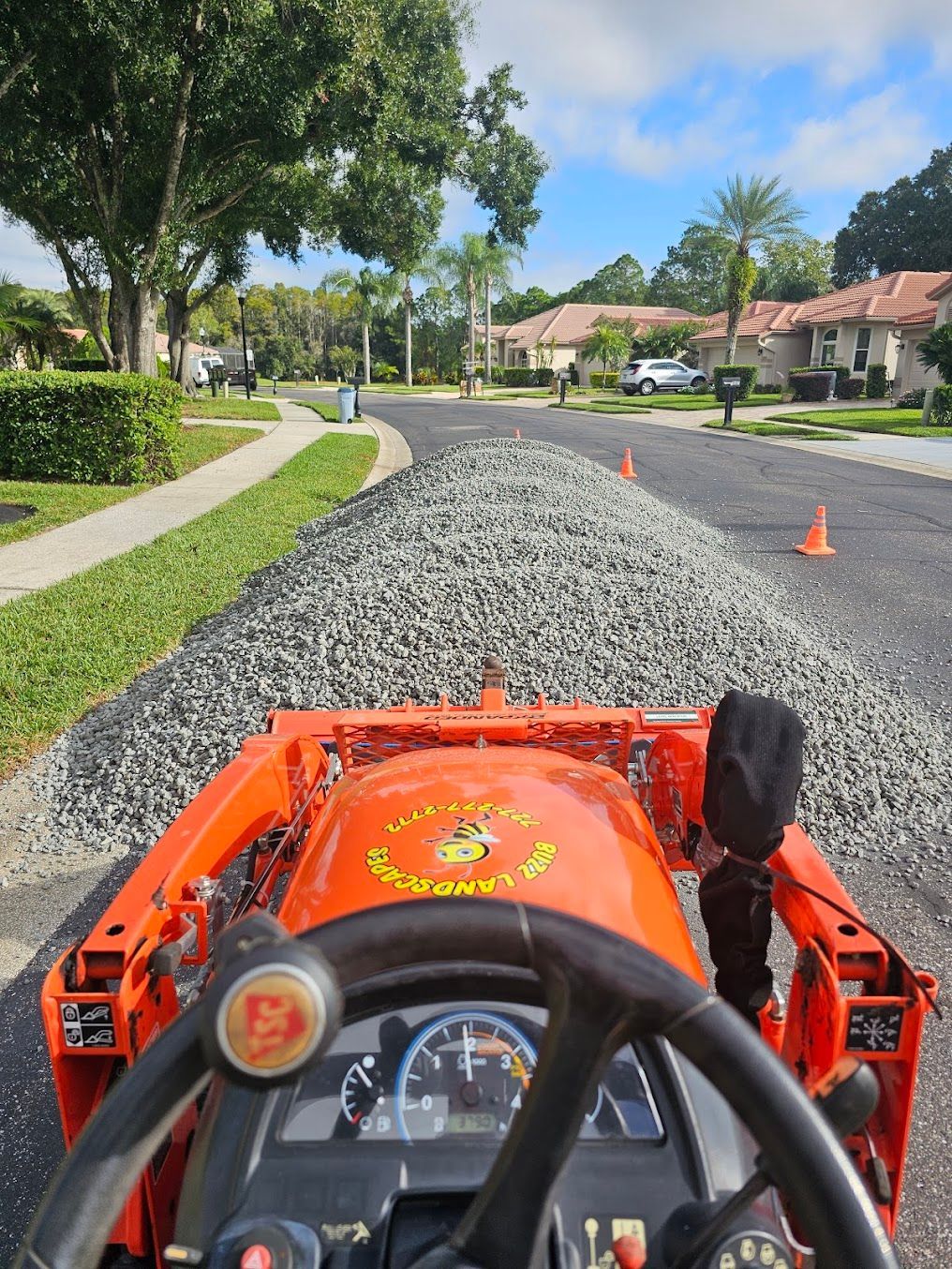 View from a ride-on mower on a gravel path in a suburban neighborhood, with cones ahead.