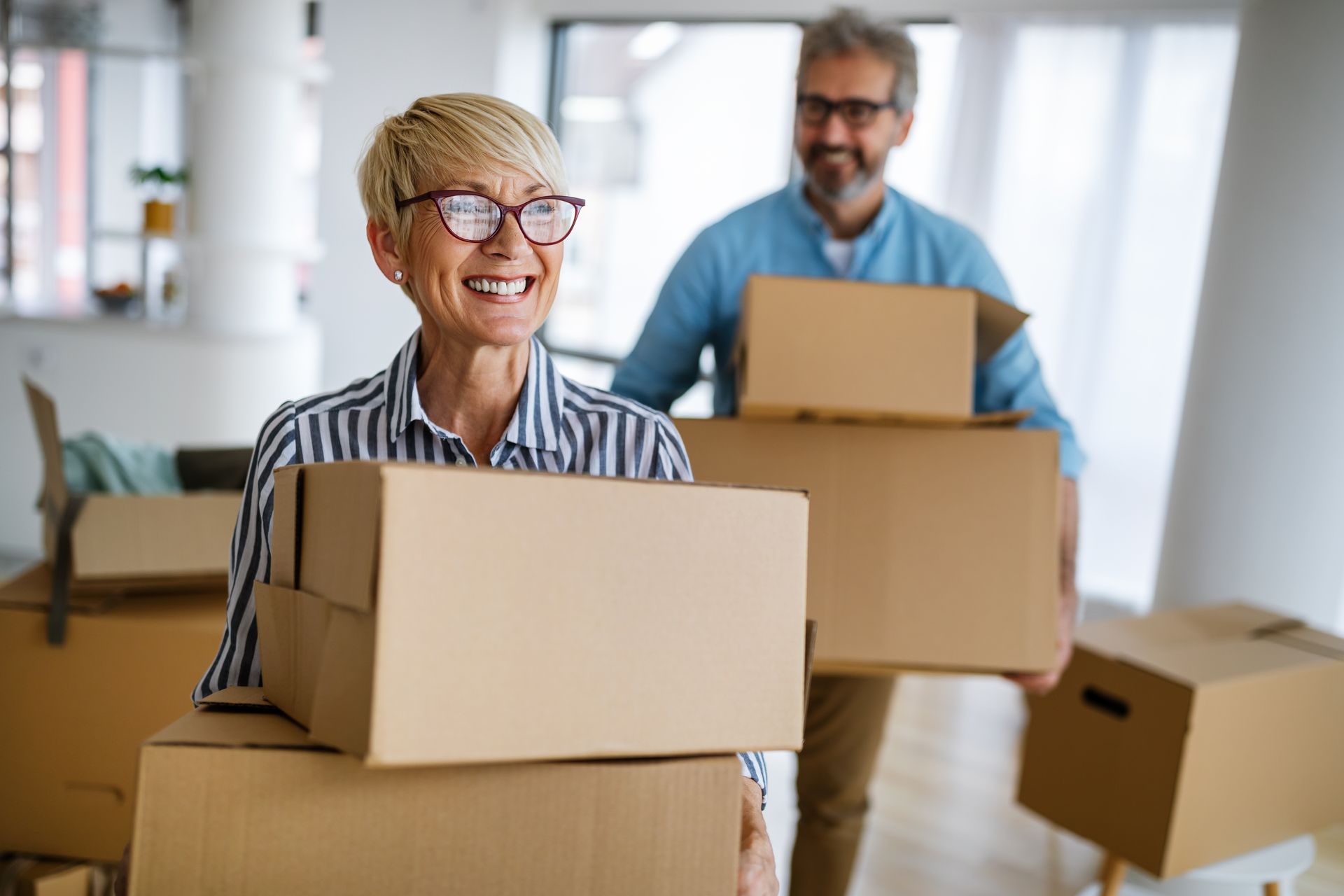 Two people carrying cardboard boxes in a bright room, smiling during a move.