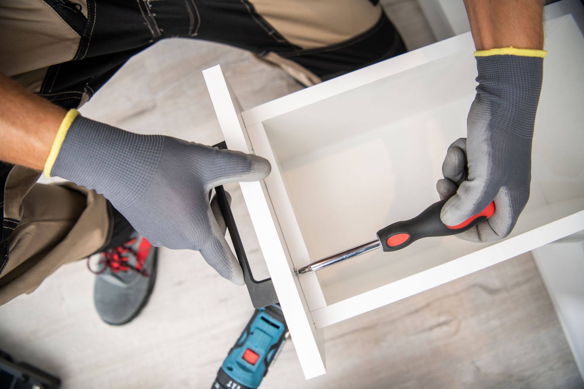 Gloved hands using a screwdriver to assemble a white cabinet frame on the floor