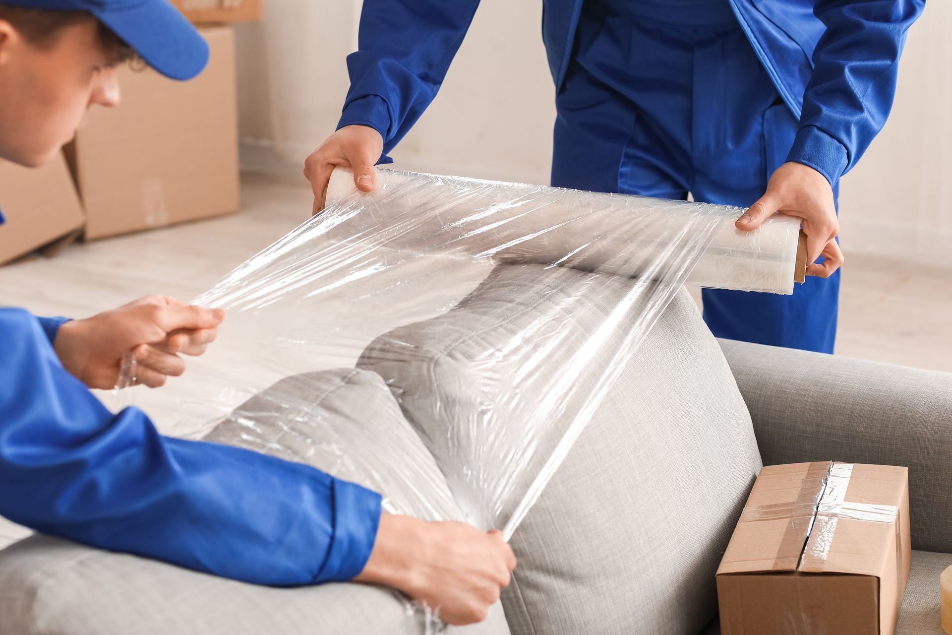 Two workers in blue uniforms wrapping a sofa in clear plastic for moving.