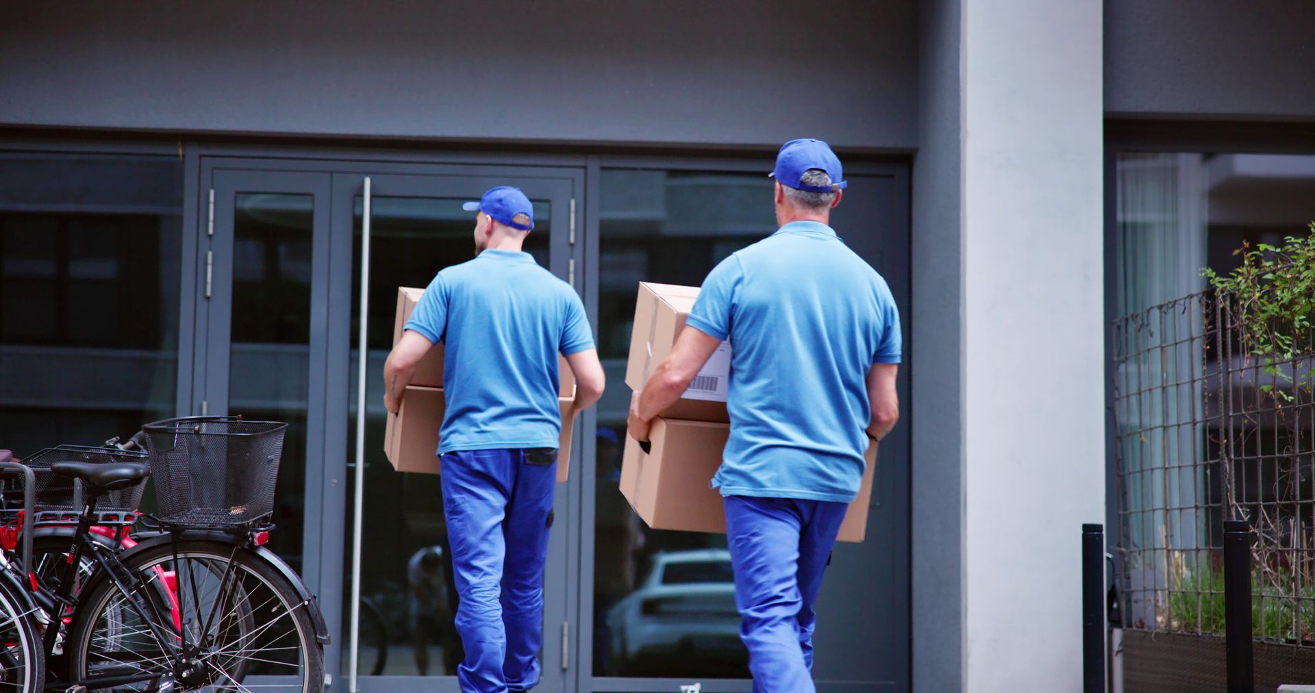 Two movers in blue uniforms carrying a cardboard box into a building entrance