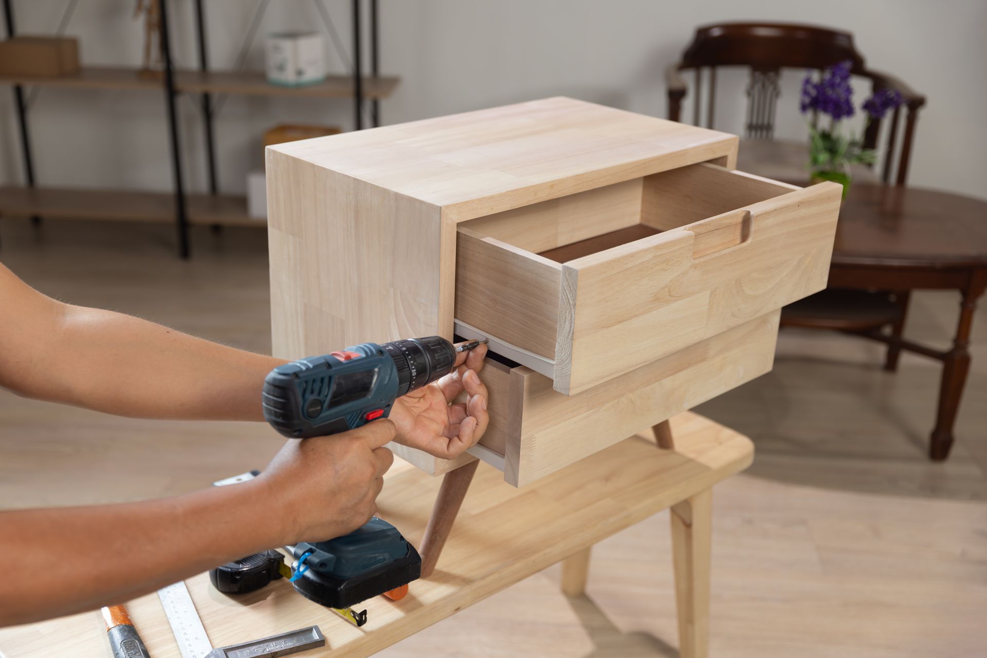 Hands assembling a light wood drawer cabinet with a power drill in a workshop.