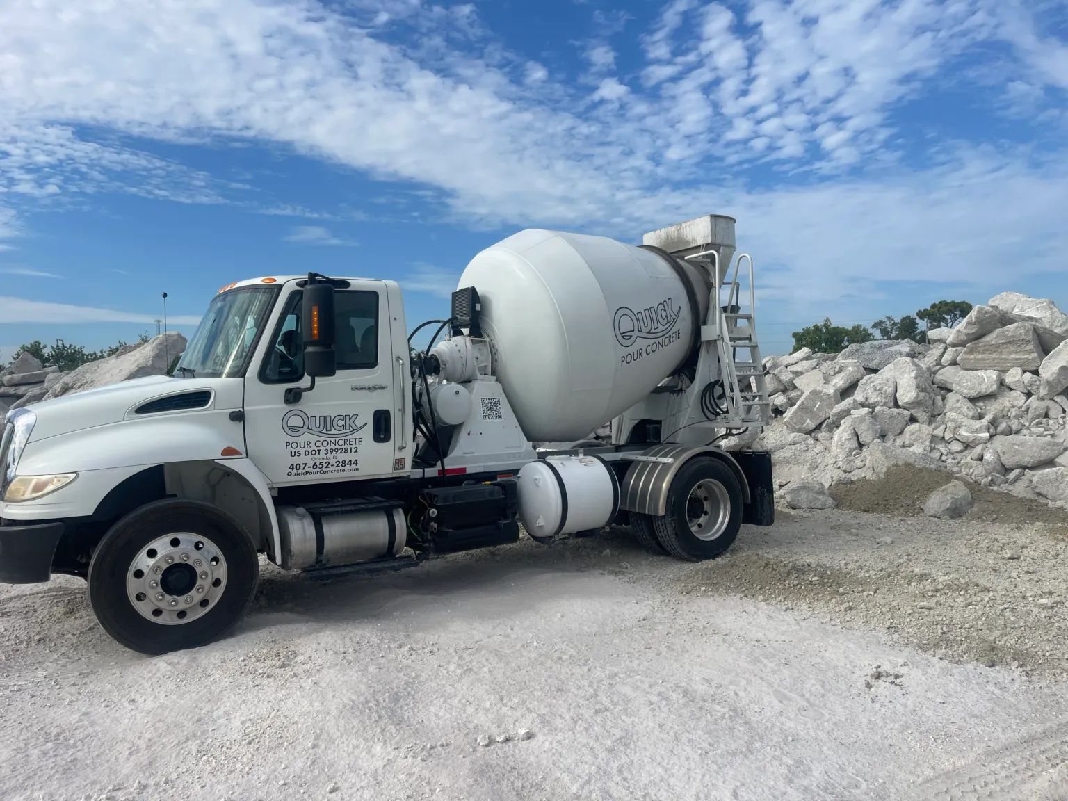 White concrete mixer truck parked on gravel, cloudy sky in background.