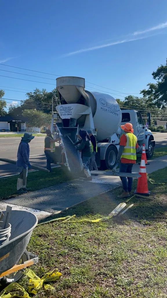 Concrete truck pouring concrete on a sidewalk; workers in safety vests; orange traffic cones.
