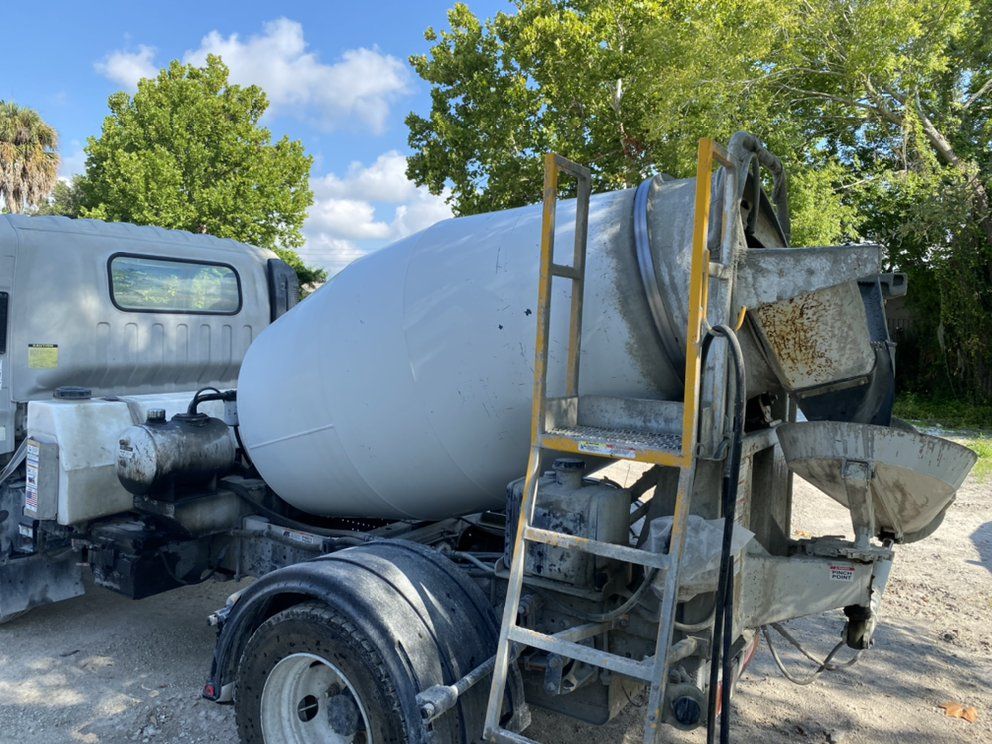 Cement mixer truck on a dirt road, with a large gray drum, yellow ladder, and trees in the background.