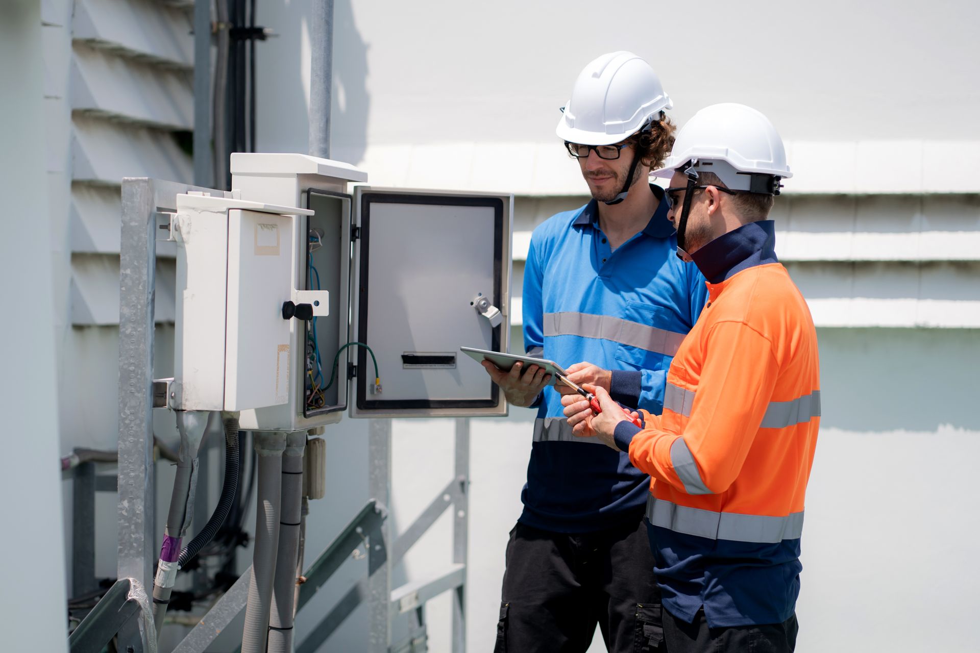 Two workers in hard hats inspecting an open electrical panel outside a building