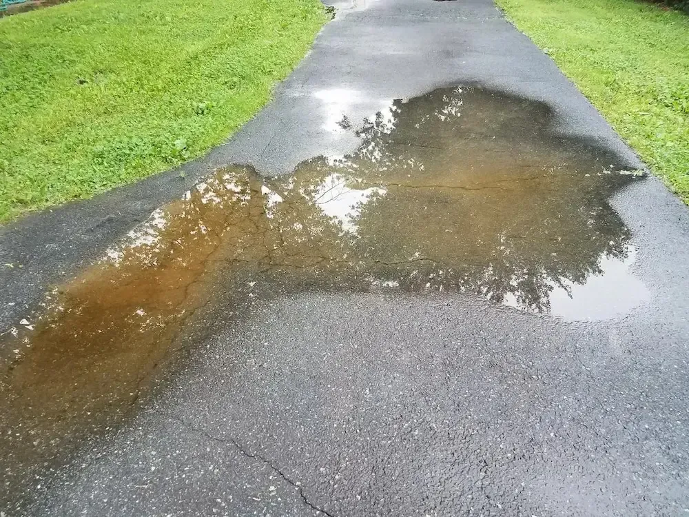 Rain puddles and brown runoff on a wet paved path beside green grass.
