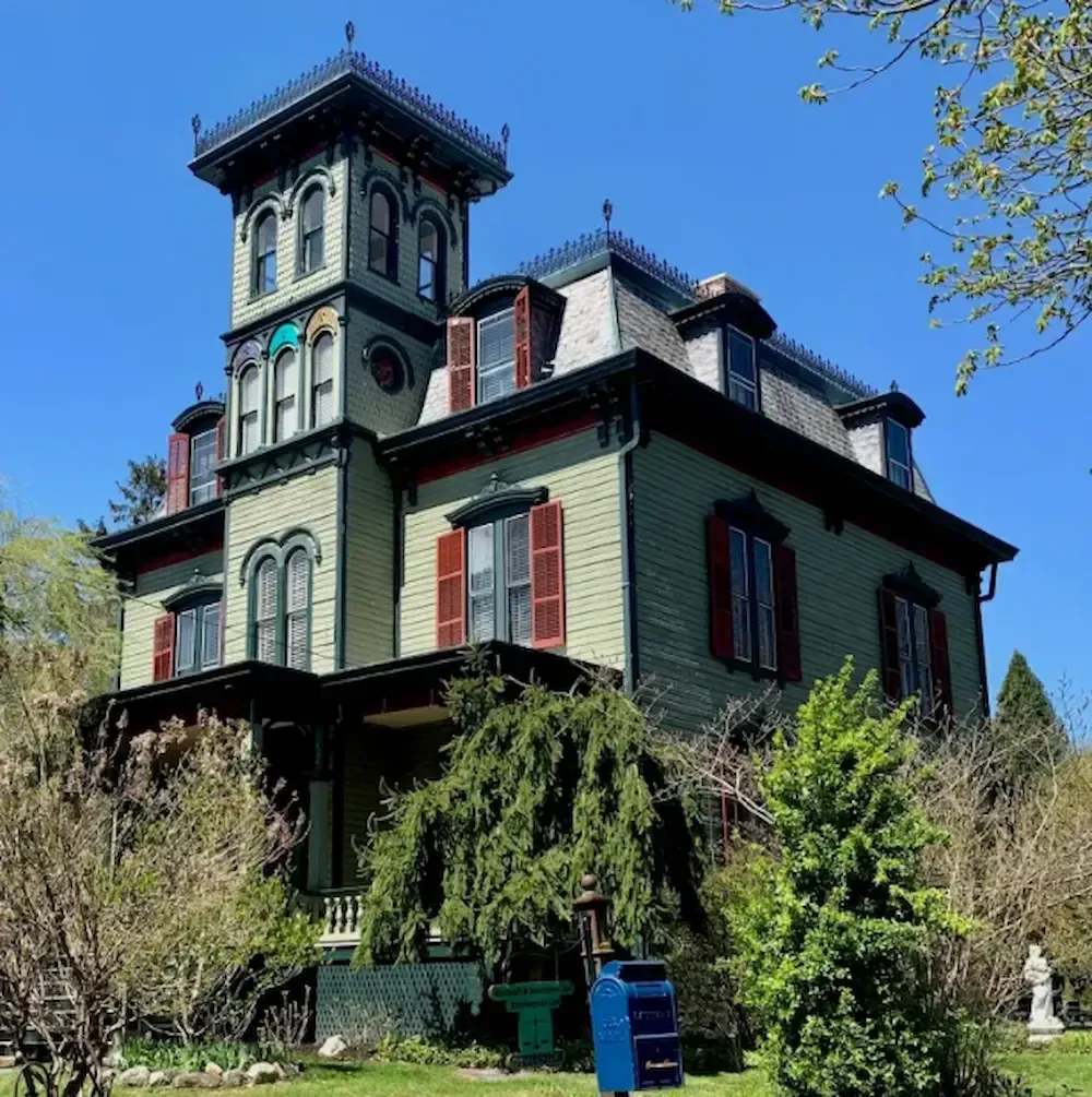 Victorian house with tower, green siding, red shutters, and overgrown garden under a blue sky