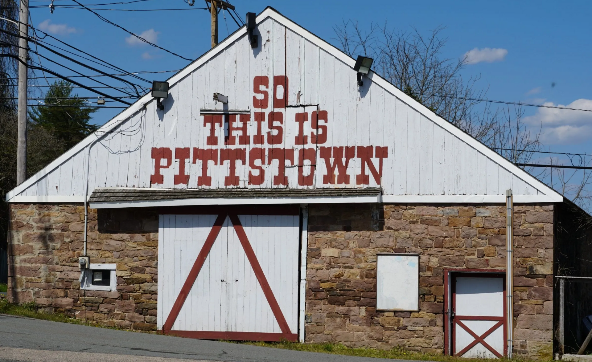Rustic barn with red “SO THIS IS PITTS TOWN” sign on a sunny street corner