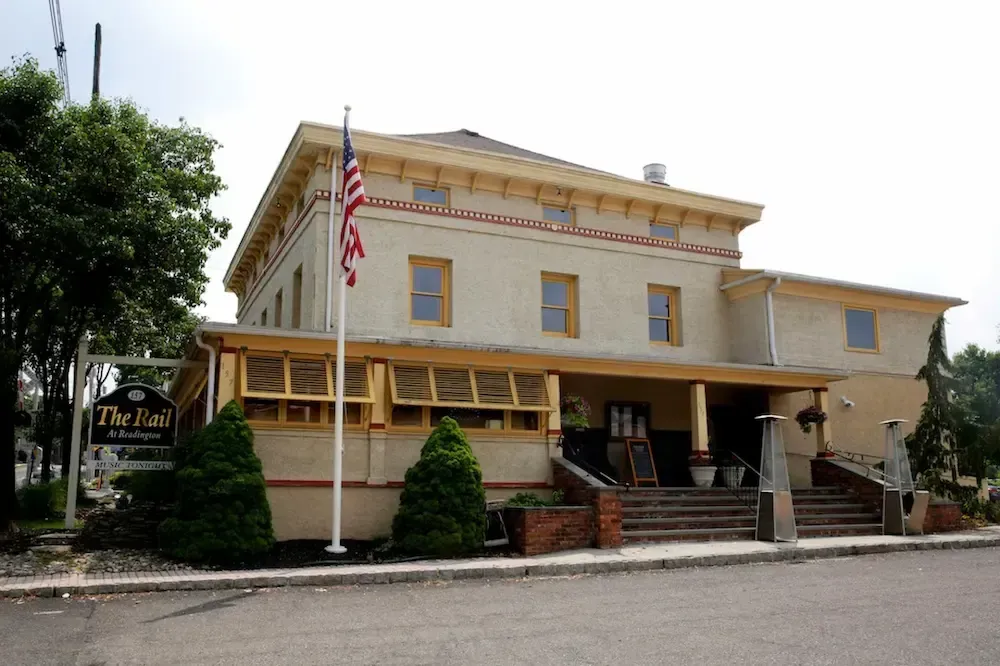 Two-story beige hotel with a red roof, porch, and American flag, beside a tree-lined street