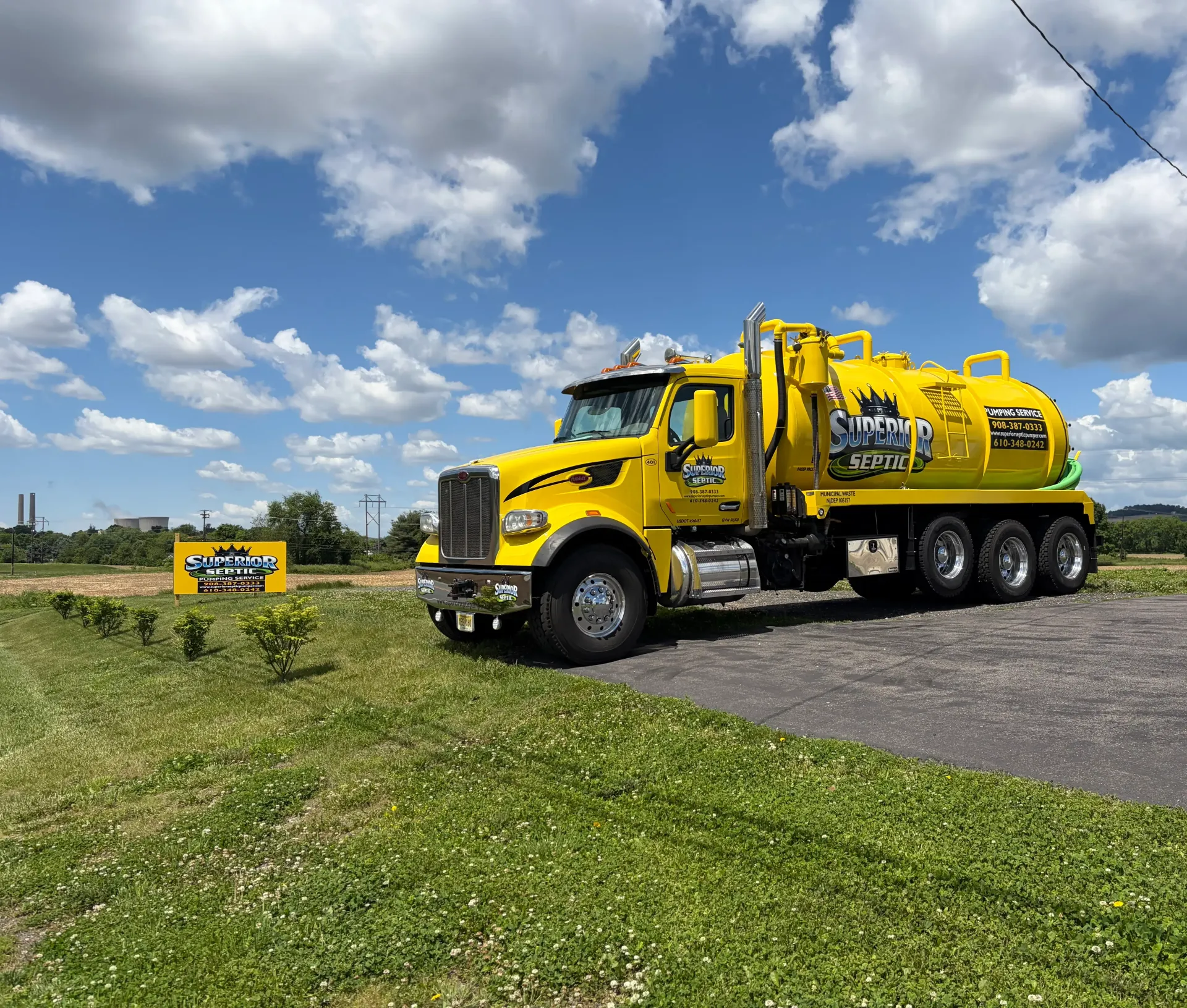 Yellow vacuum truck parked on a roadside under a blue sky with clouds, with a smaller truck in the distance.