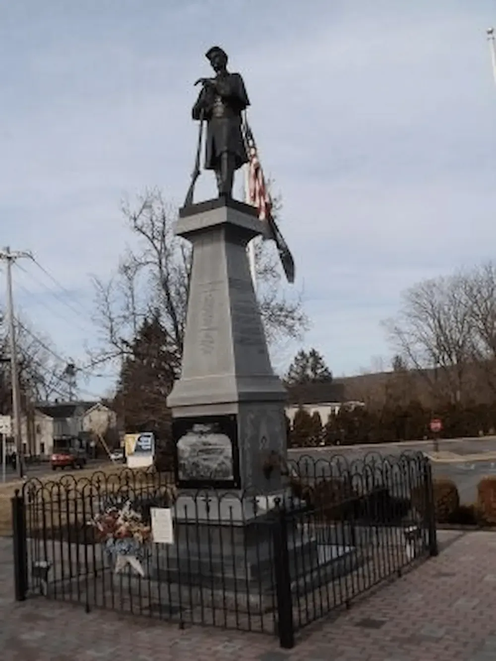Statue on a tall stone pedestal in a fenced memorial, with an American flag and neighborhood houses nearby