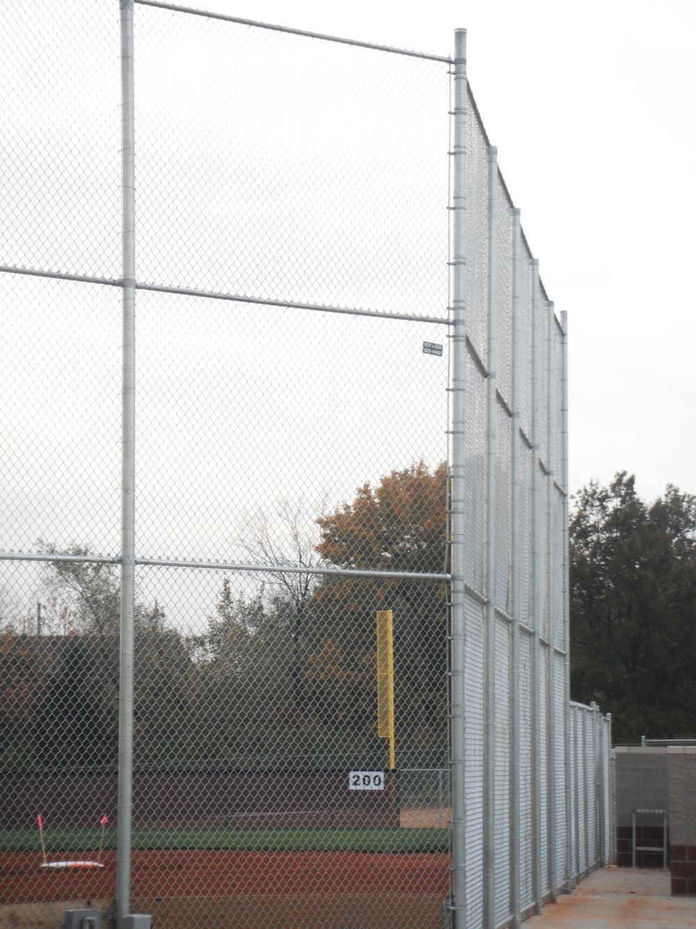 Chain-link baseball field fence. Gray metal posts and net. Red dirt infield, green grass, cloudy sky.