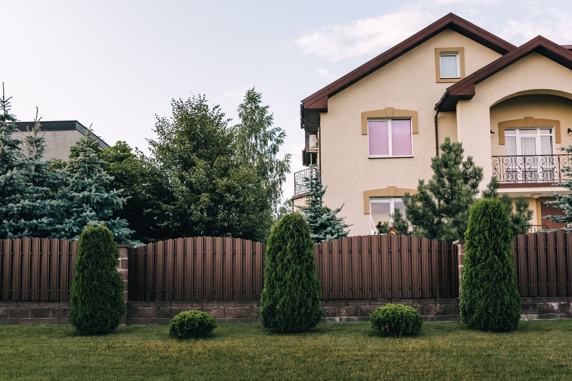 Tan house with a brown roof and fence, featuring green shrubs and trees in a manicured lawn.