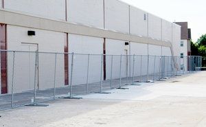 Chain-link fence in front of a light-colored building. The fence is set up on a paved surface.