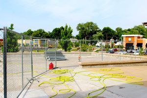 Empty, fenced-off outdoor pool with yellow hoses, likely under construction, surrounded by buildings and trees.