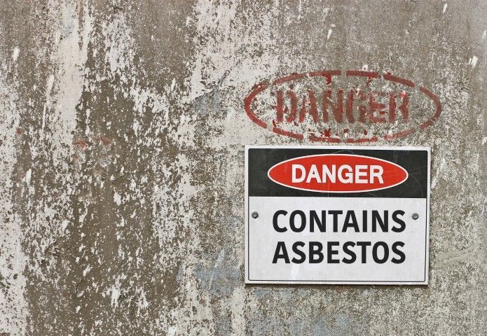 Danger sign on a wall reading “Contains Asbestos” with faded red text above.