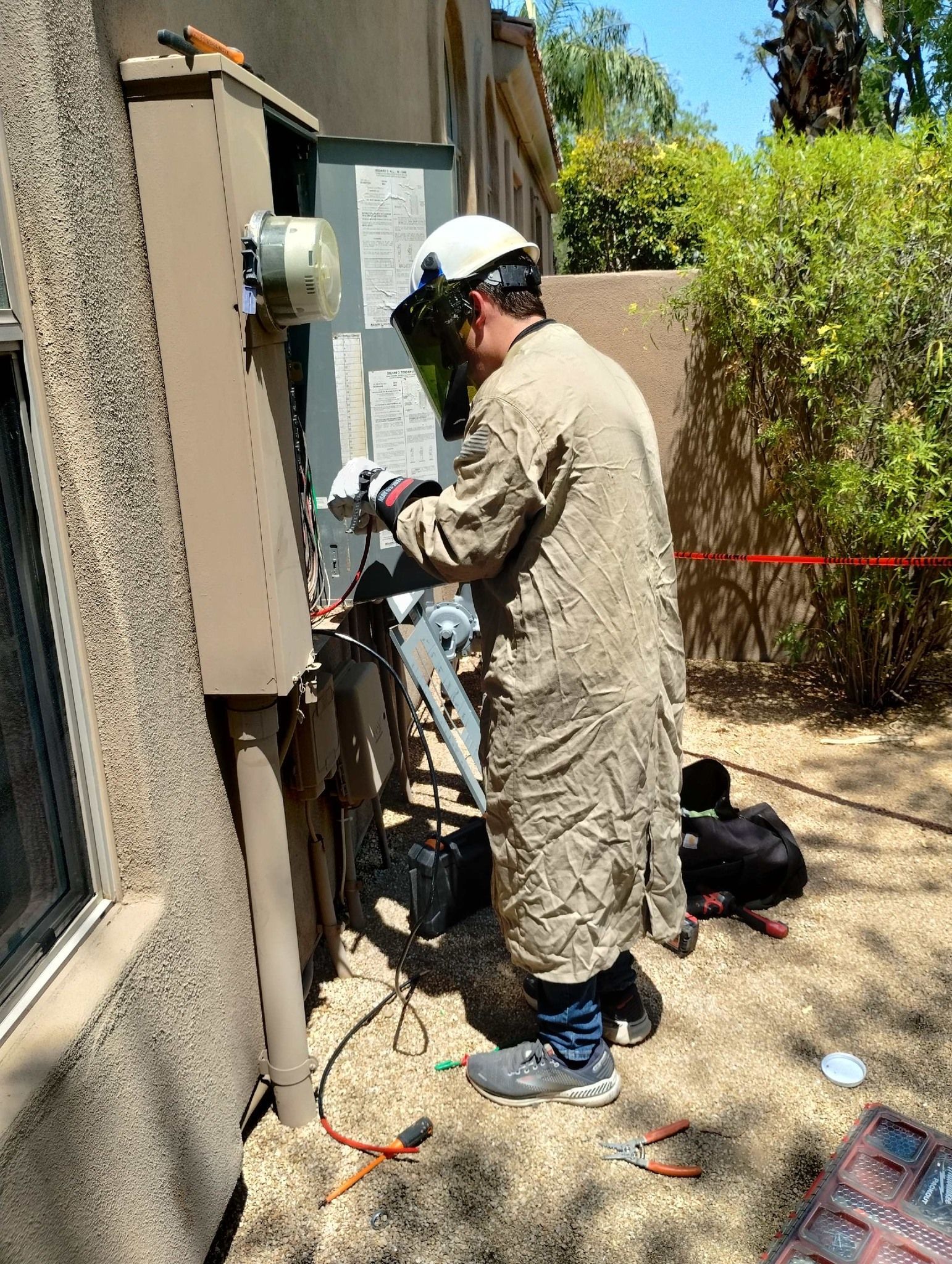 A utility worker in protective gear stands outside, working on an open electrical box attached to a stucco wall.