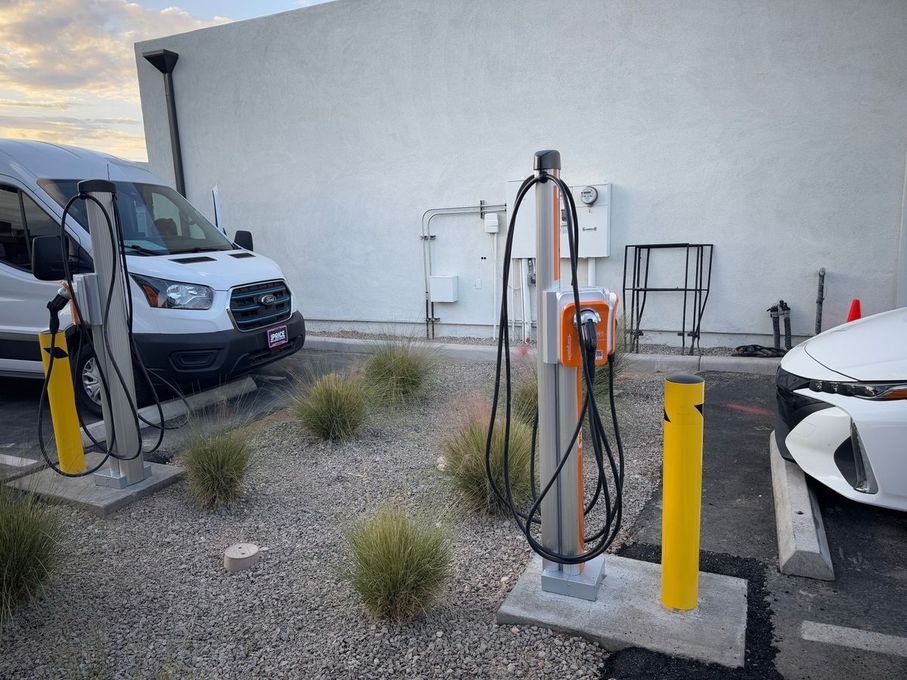 Two electric vehicle charging stations installed on concrete pads in a gravel lot with cars parked nearby.