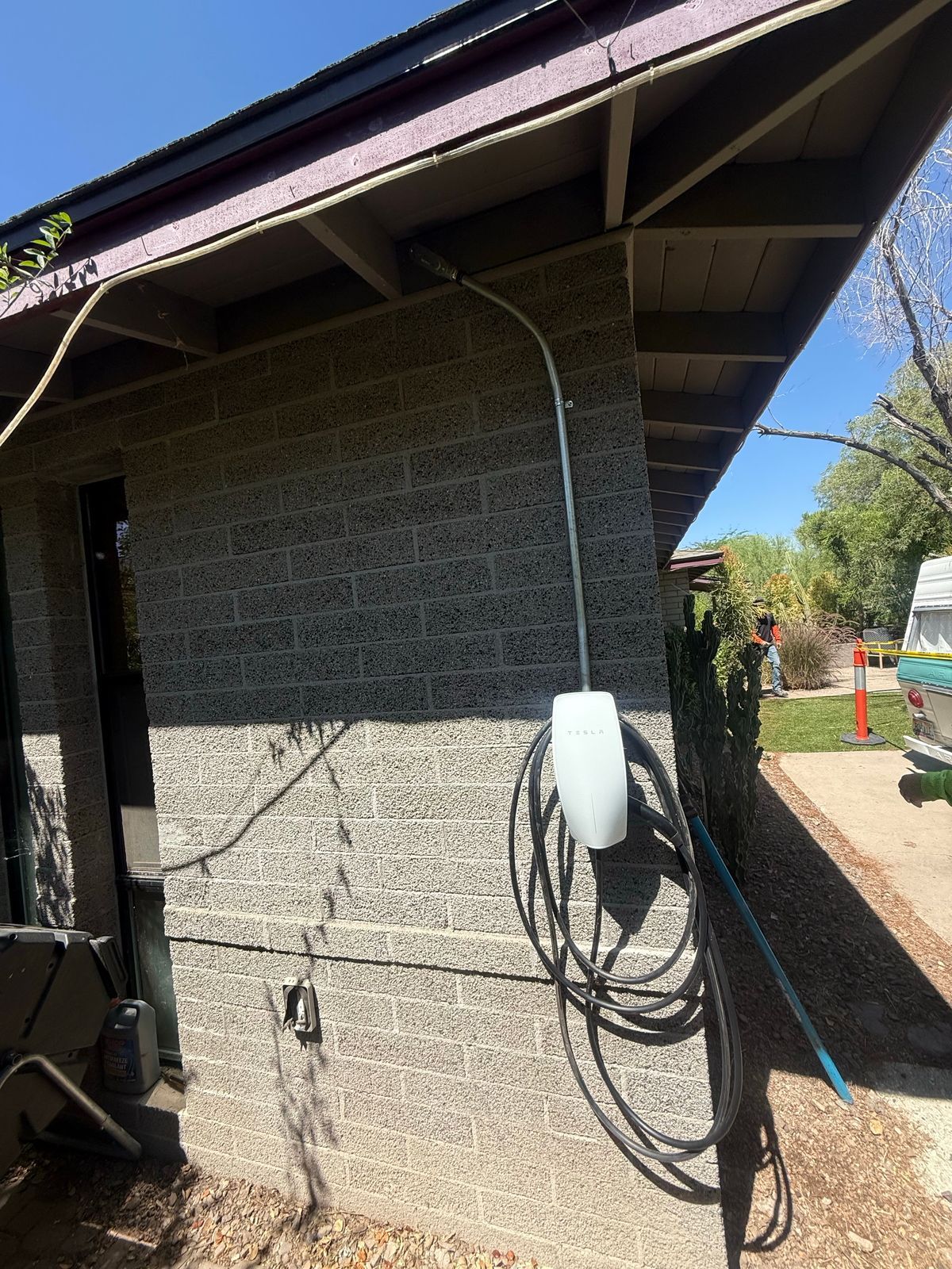 A white Tesla electric vehicle wall charger mounted on a gray concrete block wall under a roof overhang.