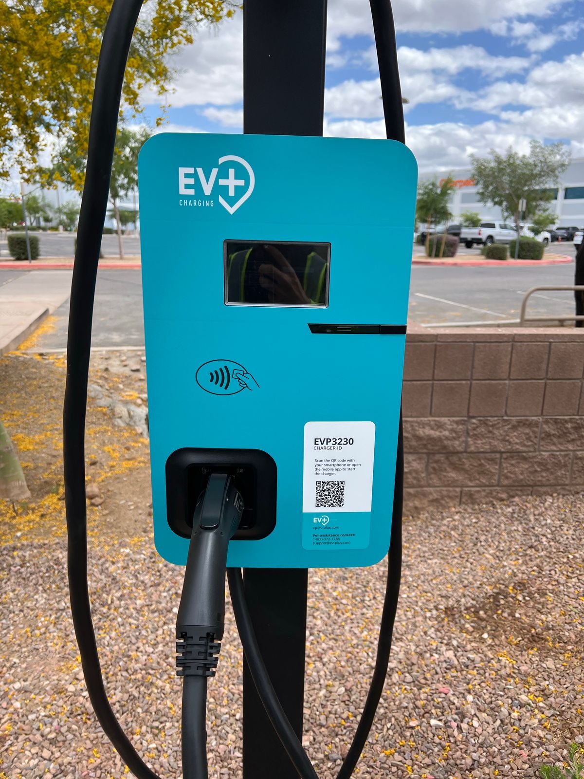 A teal EV+ electric vehicle charging station mounted on a post in an outdoor parking lot with a charging cable attached.