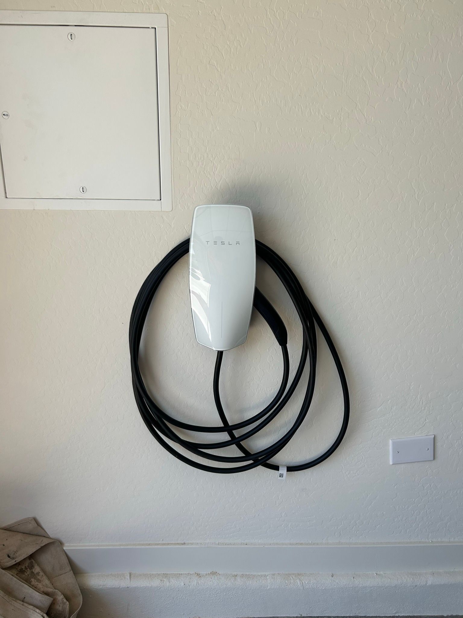 A white Tesla Wall Connector electric vehicle charger mounted on a textured interior wall with its cable coiled around it.