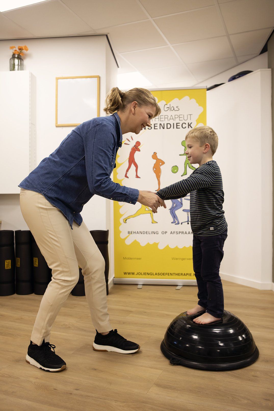 Vrouw helpt lachend een kind op een evenwichtsbal in een therapieruimte. Banner op de achtergrond.