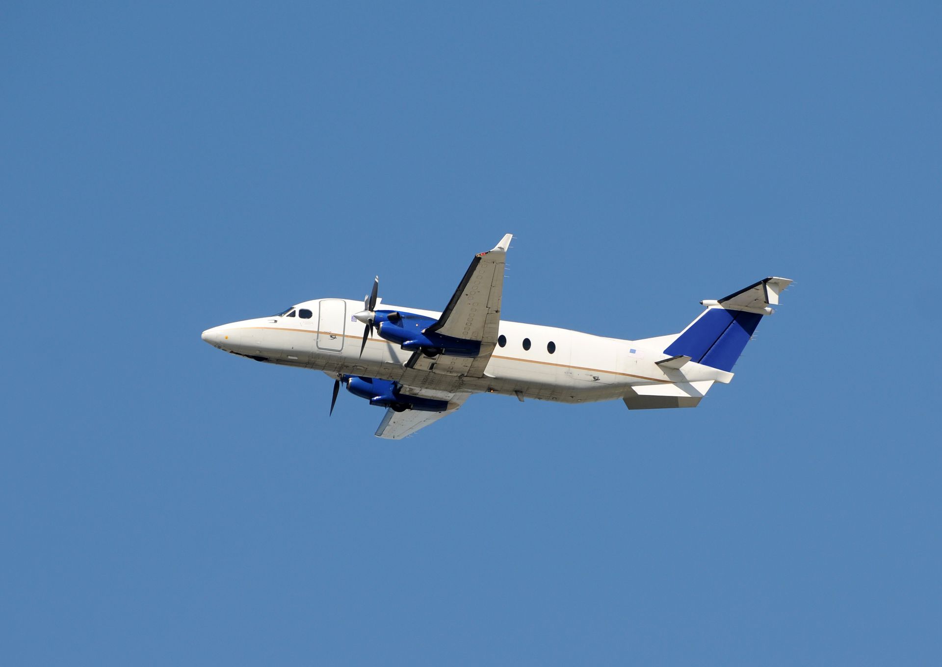 White and blue twin-engine airplane in flight against a clear blue sky.
