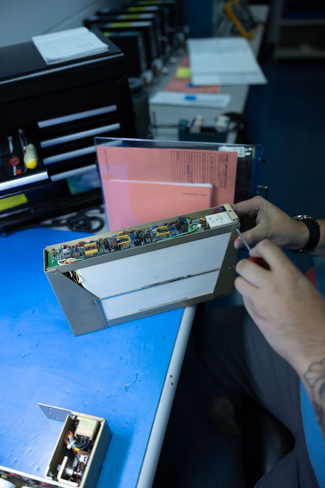 Person repairing electronic device on a blue table. They use a screwdriver, with other devices and papers in the background.