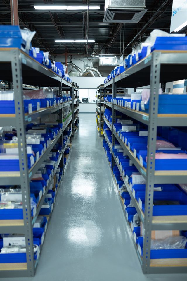 Rows of shelves filled with blue bins in a warehouse setting; reflective floor; overhead lighting.